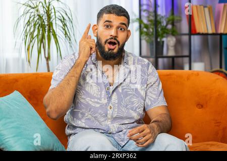 Eureka. Thoughtful clever inspired Indian man make gesture raises finger came up with creative plan feels excited with good idea inspiration motivation, question at home apartment. Guy sits on couch Stock Photo