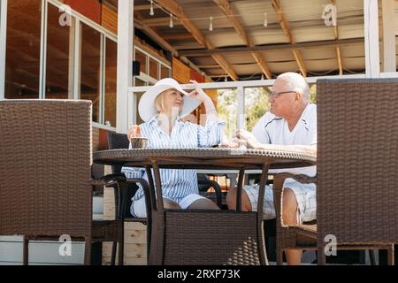 Happy mature middle aged couple talking, woman showing off her hat, flirting. On a date in a cafe. Stock Photo