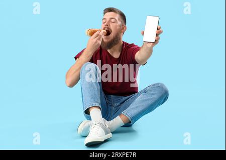 Happy young man eating tasty hot dog and showing smartphone with empty screen while sitting on blue background Stock Photo