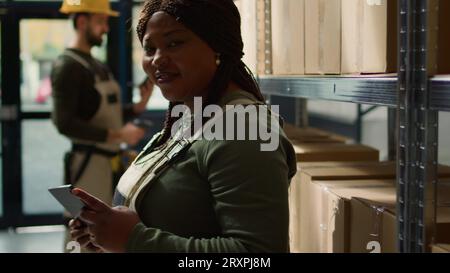 Portrait of smiling warehouse head of logistics holding digital tablet, checking packaging, ensuring flawless operations while employee in background handles packaged merchandise in cardboard boxes Stock Photo