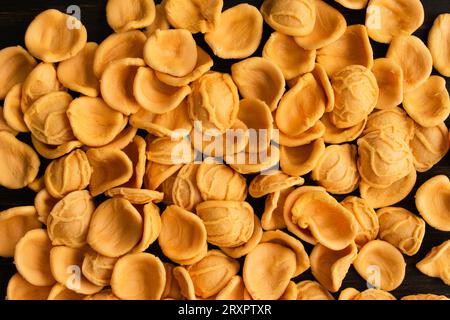 Dried Orange orecchiette Pasta Scatttered on a Wooden Table: Pieces of uncooked orecchiette pasta on a dark wood background Stock Photo