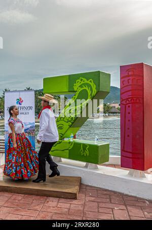 Zihuatanejo, Mexico - July 18, 2023: Young female-male couple, colorfully dressesd, performs tradional folk dance on harbor pier against first 2 giant Stock Photo