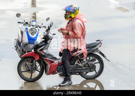 SAMUT PRAKAN, THAILAND, SEP 20 2023, A man in a raincoat sits on a motorcycle and holds a mobile phone Stock Photo