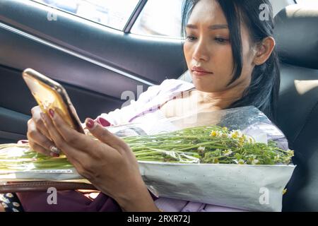 Young woman with flowers is watching her mobile phone while sitting in the car Stock Photo