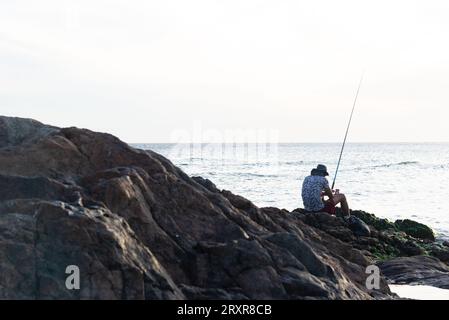 Salvador, Bahia, Brazil - December 31, 2021: Silhouette of a fisherman with fishing rod sitting on the rocks of the Rio Vermelho beach in the city of Stock Photo