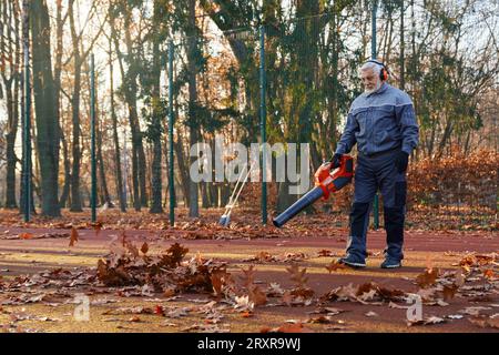 Senior man in overalls, headphones and gloves cleaning fenced city park area from dry fall leaves. Side view of professional janitor using hands blower during working process outside. Work concept. Stock Photo
