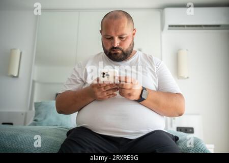 Fat bearded bald guy with smartphone chatting, dials number, types message, watches news, weather Stock Photo