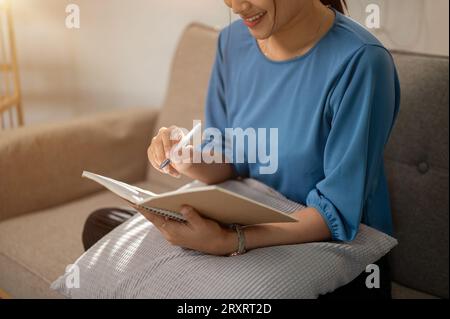 A close-up image of an attractive Asian woman in casual clothes sitting on a sofa and making her grocery list in a book or writing something in her no Stock Photo