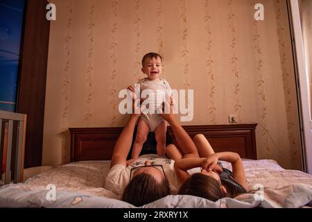 Diversified father in glasses throws baby son on the bed, eldest daughter is having fun nearby. Family fools around and plays on weekends. Father's da Stock Photo