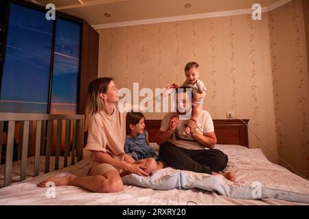 Funny portrait of diverse cheerful family on bed in morning. Stylish mother hugs daughter, father with glasses holds little son on shoulders. Parents Stock Photo