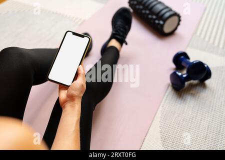 Female sports person sits on yoga mat and holding smart phone with white empty screen in her hand. Mobile phone and fitness. Workout mobile app. Stock Photo