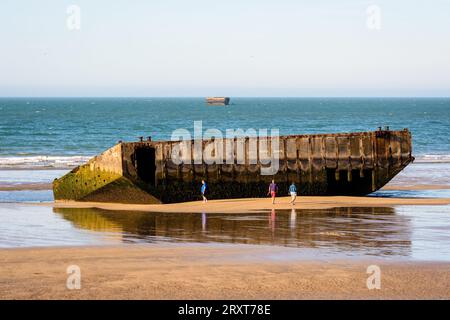 People stroll on the beach next to remains of the Mulberry harbor built on Gold Beach in Arromanches, France, after the Normandy landings in WWII. Stock Photo