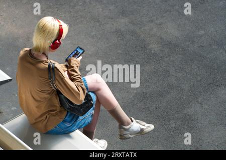 Young person looking at phone wearing headphones and sneakers sitting on seat using social media, in Melbourne city, Australia. Stock Photo