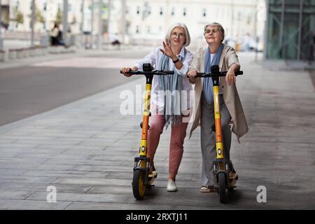 Senior women with gray hair enjoying ride together on electric scooters outdoors on streets. Elderly female friends talking and travel around city. Stock Photo