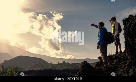 A mature couple with backpacks, wearing windbreakers, stand atop a rock, shielding their eyes from the sun as they take in the panoramic view. The exp Stock Photo