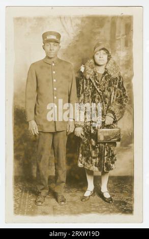 Black-and-white photographic postcard that was never stamped and mailed but has written in pencil on the back the name 'Omer Ester'. The image on the postcard is of Pullman Porter, Omer Ester, in his full porter uniform, including Pullman Porter cap. Standing next to him is an unidentified African American woman wearing what appears to be a velvet full length coat with fur trim on the collar and sleeves. She is wearing a 1920s style woman's hat and holding a handbag in her right hand. They are in a photography studio standing in front of a painted backdrop with an oriental-style carpet under t Stock Photo