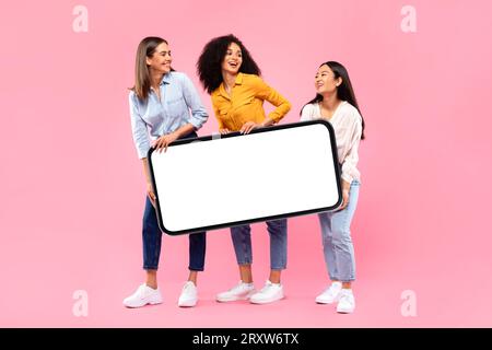 Happy young multiethnic girlfriends carrying huge cellphone with blank screen, posing on pink background, full length Stock Photo