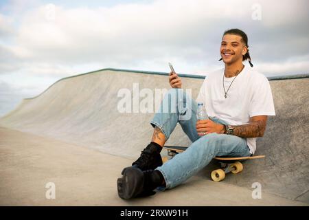 Happy european man using mobile phone while sitting on skateboard on sports ramp, guy smiling at camera, free space Stock Photo