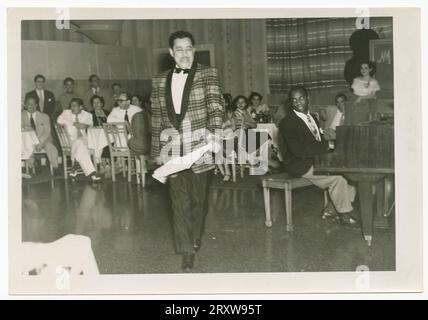 B & W photograph of Cab Calloway dancing while a man plays the piano. Calloway is holding a white handkerchief in his PL hand, and is depicted wearing a plaid suit jacket with dark pants. A large group of people are seat at tables behind Calloway. A yellow sticky note attached to back of photograph reads, [Hotel/National/in/Cuba]. Stock Photo