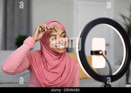Muslim beauty blogger lady making facial massage using stone roller and making video review on cellphone for online blog Stock Photo