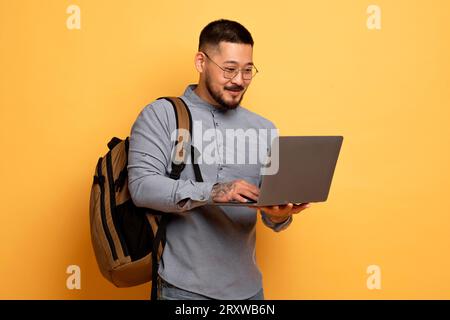 Young Asian Student Guy With Laptop And Backpack Standing On Yellow Background Stock Photo