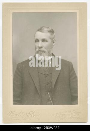 A black-and-white photograph of a man photographed at the Ball & Sons studio of Seattle, WA. The photo is mounted on a light colored cabinet card stamped with the Ball & Sons logo and store location on the bottom of the front side. The man is seated and photographed from the waist up. He wears a dark suit and cravat. He has white hair and a full beard and looks off to his right in the photo. On the back of the card mount there is the impression of an oval photograph of a young boy. Stock Photo