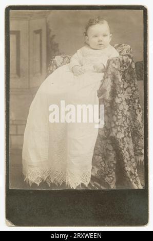 A black-and-white photograph of a baby. The photo is mounted on a dark colored cabinet card with no writing on the front or back. The child has curly hair and is dressed in a long white gown. He or she is placed in a textile covered chair and positioned in front of a painted backdrop. Stock Photo