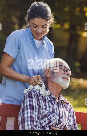 Young woman nurse massaging senior man on back and shoulder in park Stock Photo
