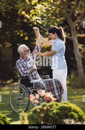 Young woman physiotherapist helping senior man in wheelchair with stretching painful hand by doing exercises in park Stock Photo