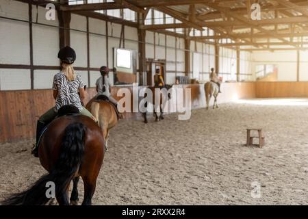 Horse riding school. Little children girls at group training equestrian lessons in indoor ranch horse riding hall. Cute little beginner blond girl kid Stock Photo