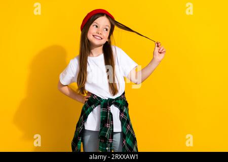 Photo portrait of adorable small girl look empty space hold strand hair dressed stylish white outfit isolated on yellow color background Stock Photo