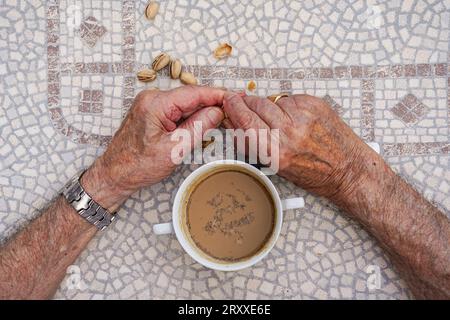 senior man wrinkled hand drinking a coffee and eating pistachios. active old age making a snack. Aerial view. Stock Photo
