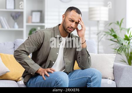 A young African-American man is suffering from headaches, migraines, and dizziness. Sits at home on the sofa and grimacing, holding his head. Stock Photo
