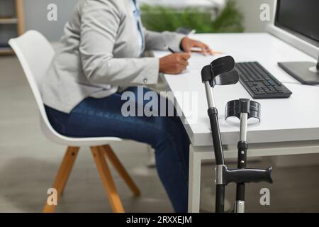 Crutches by an office desk with a disabled woman working on a computer in the background Stock Photo