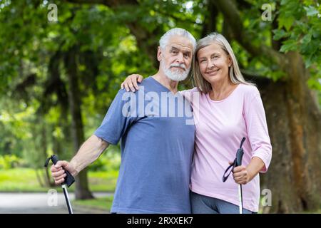 Portrait of a gray-haired senior man and woman standing outside in a park hugging each other and holding trekking poles. They go on a Norwegian walk together. Stock Photo