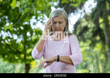 An older gray-haired woman feels bad after jogging and active sports. Standing in the park, holding his head and looking at the smart watch, checking his pulse and breathing. Stock Photo