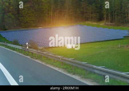 Solar panels of a power plant in a clearing near the forest in the evening, against the backdrop of the setting sun. Stock Photo