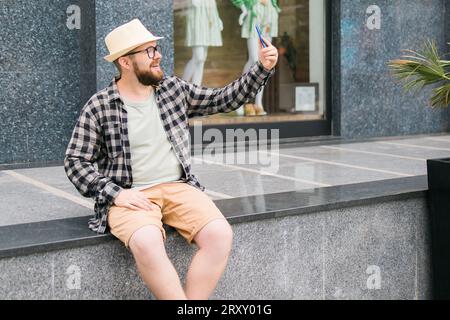 Bearded millennial man takes selfie via smartphone strolls in downtown poses against modern city building enjoys leisure time copy space. People urban Stock Photo