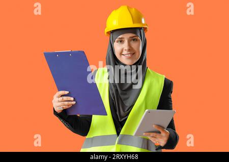 Female Muslim construction worker with clipboard and tablet computer on orange background Stock Photo
