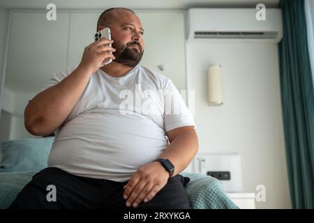 Fat guy calls on phone, sitting on bed, looking out window, signs up for gym to put figure in order Stock Photo