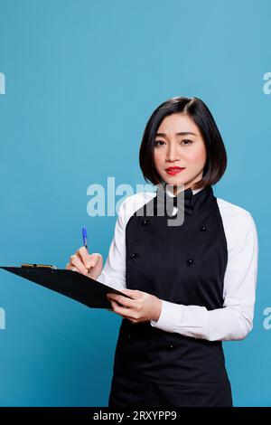 Attractive asian waitress taking notes of customer order in restaurant portrait. Young woman receptionist maintaining records, writing on clipboard with pen and looking at camera Stock Photo