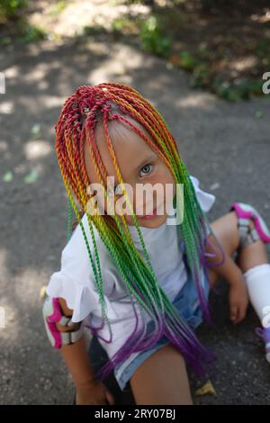 little cute girl with colorful afro pigtails sits on a skateboard Stock Photo