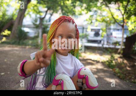 little cute girl with colorful afro pigtails sits on a skateboard and shows a peace sign Stock Photo