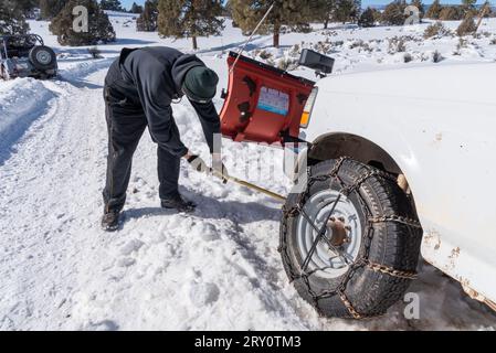 Male bends over to shovel snow from beneath a pickup truck, tires with chains, that has slid off the road into the ditch, Lumberton, New Mexico, USA. Stock Photo