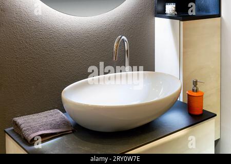 Modern white ceramic oval washbasin placed in bathroom near mirror with bright frame and towel with soap Stock Photo