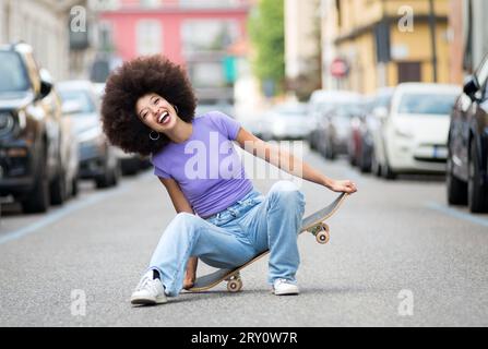 Full body of cheerful young African American female skater in casual clothes having fun on skateboard in street Stock Photo