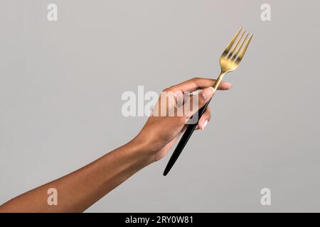 Crop anonymous hand of African American female holding gold colored fork against gray background Stock Photo