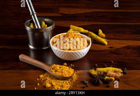 Turmeric powder kept in white bowl and wooden spoon with Mortar and Pestle, dry root of turmeric, dry spices kept around as prop on brown wooden surfa Stock Photo