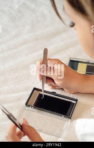 Close up of young lashmaker taking black artificial eyelash from palette using tweezers. Concentrated woman carefully looking at container of material Stock Photo