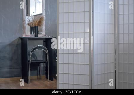 Interior of modern light room in apartment with white folding screen standing near black table with vase of pampas grass. Beige plants reflecting in m Stock Photo